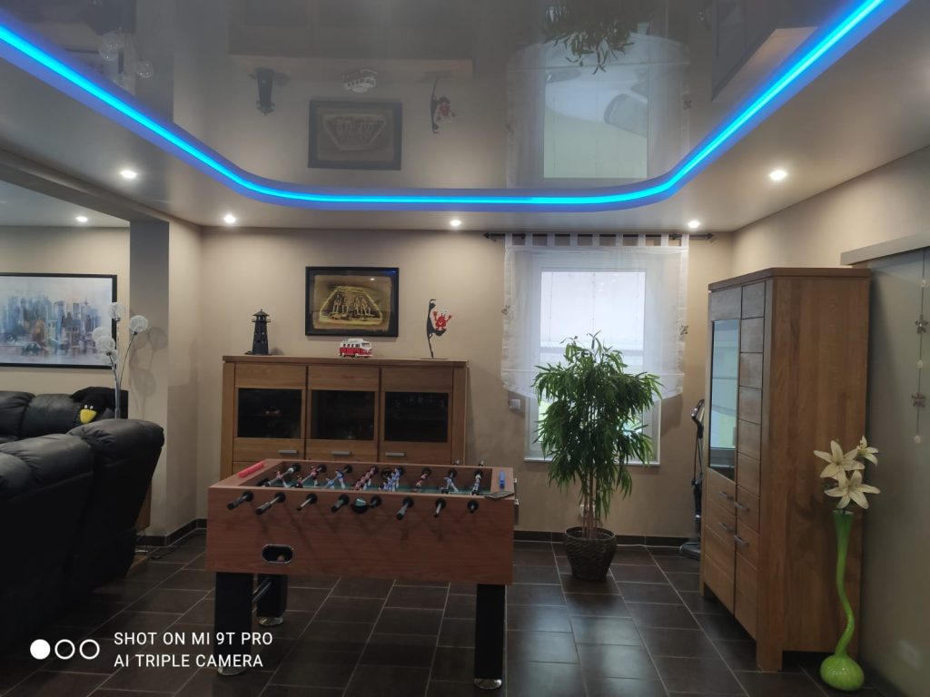 Modernes Wohnzimmer mit Hochglanz-Spanndecke, blauer LED-Lichtlinie und integrierten Spots
